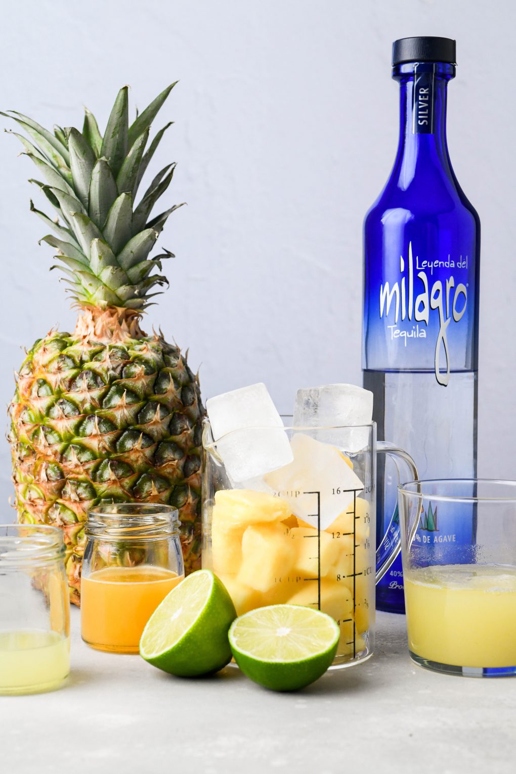 Straight on image of ingredients for pineapple margaritas - a fresh pineapple, frozen pinapple, ice cubes, fresh fruit juices, and blanco tequila. On a light colored background.