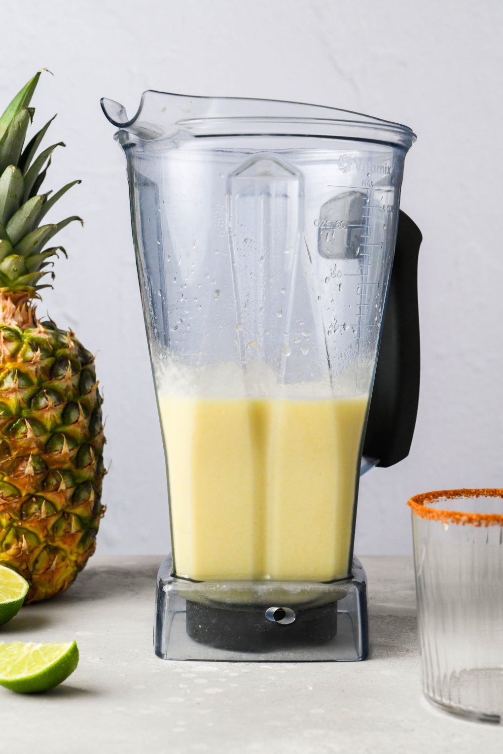 Straight on image of frozen pineapple margaritas, in a blender container - on a light colored background next to a fresh pineapple and a bottle of milagro silver tequila.