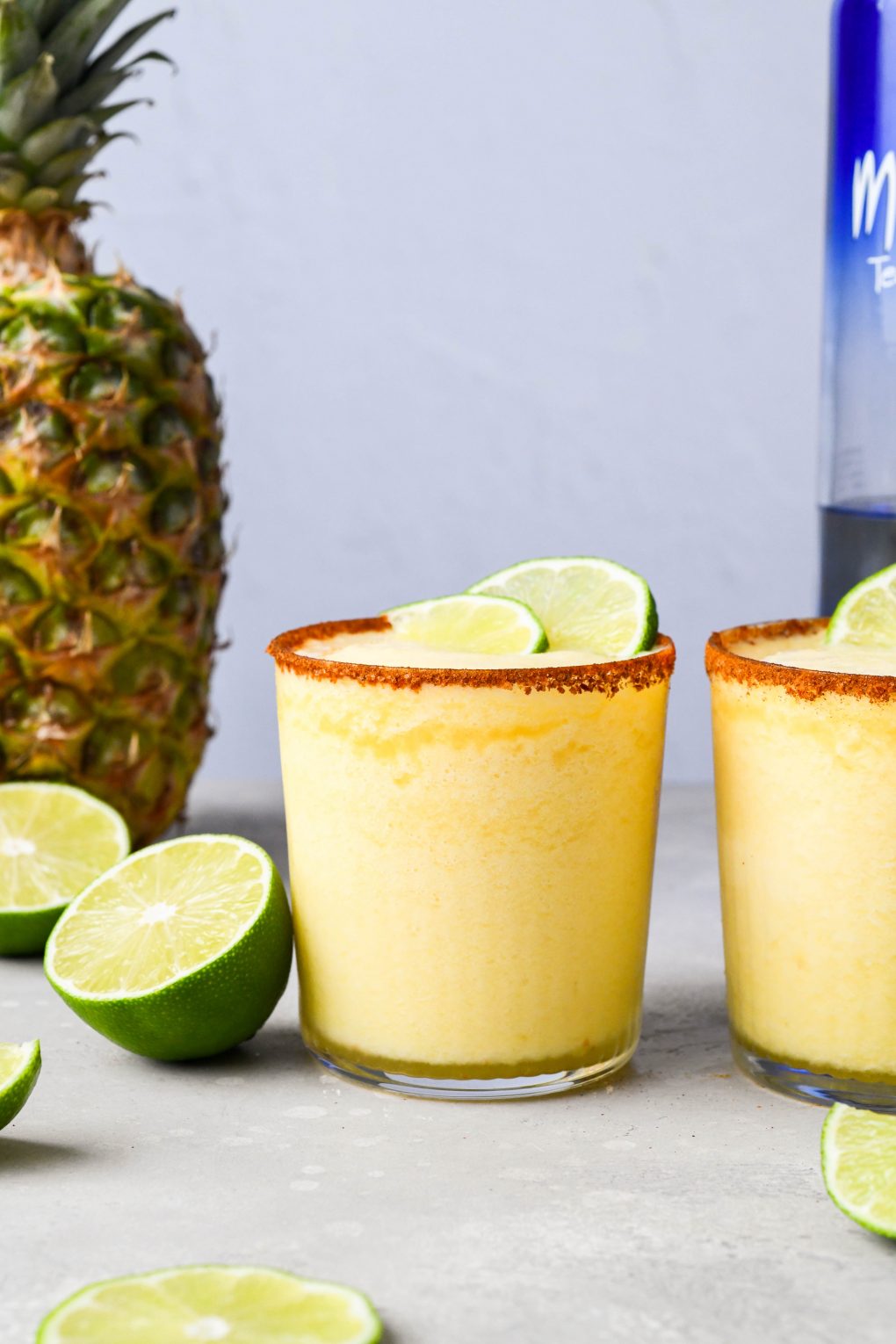 Straight on shot of two side by side frozen pineapple margaritas on a light background next to a fresh pineapple and some cut lime wedges.
