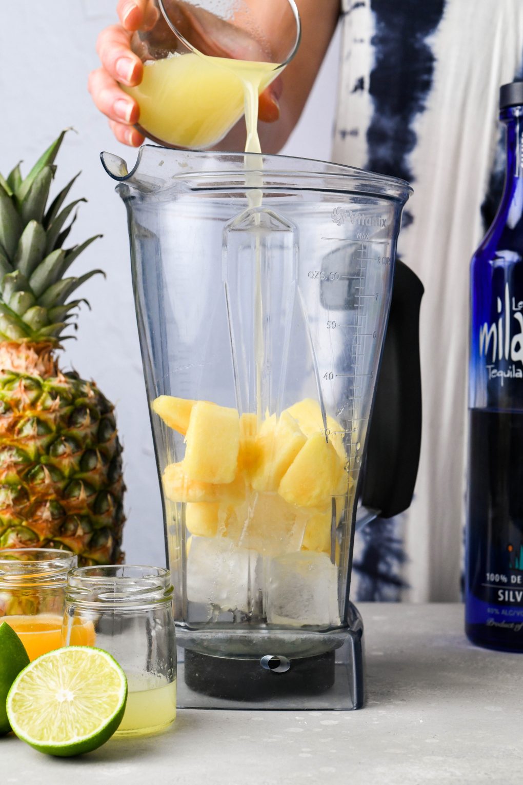 A hand pouring pineapple juice into a blender with frozen pineapple and ice. On a light background next to a fresh pineapple and a bottle of tequila.