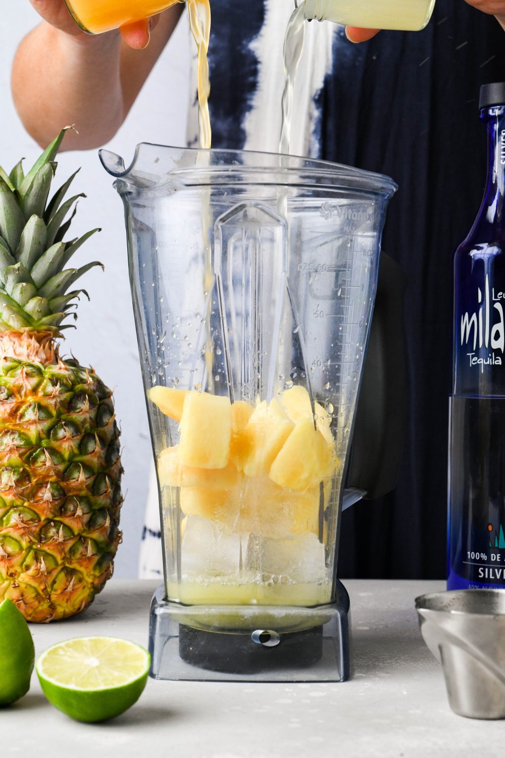 A hand pouring fresh lime and orange juice into a blender with frozen pineapple and ice. On a light background next to a fresh pineapple and a bottle of tequila.