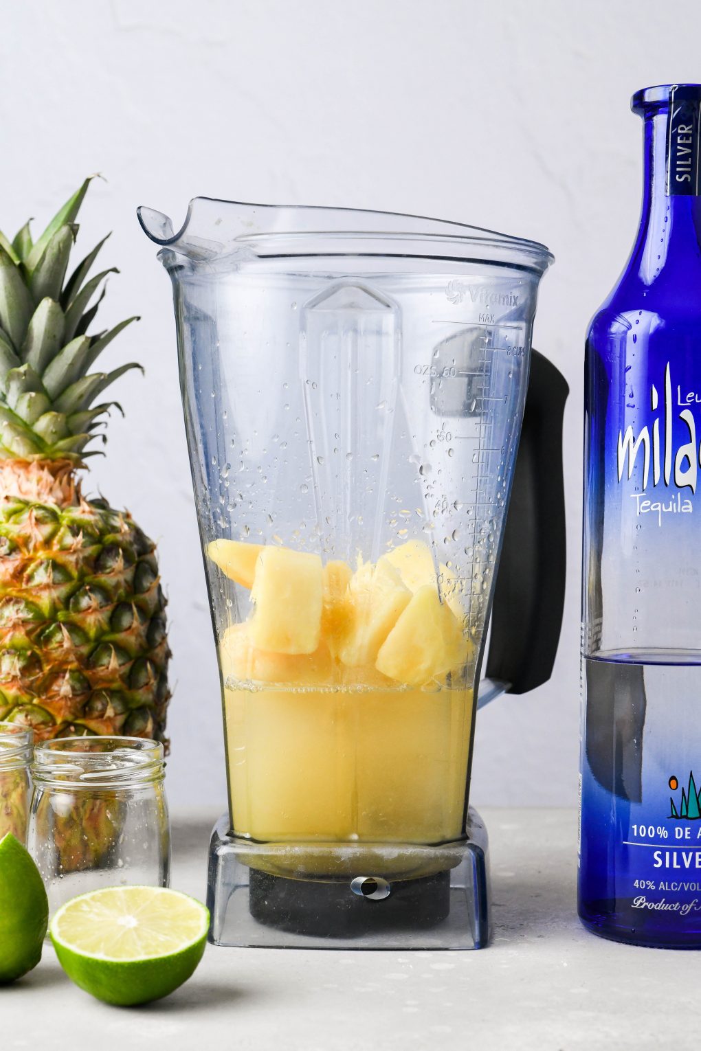Straight on image of frozen pineapple, ice, juices, and tequila in a blender container - on a light colored background next to a fresh pineapple and a bottle of milagro silver tequila.