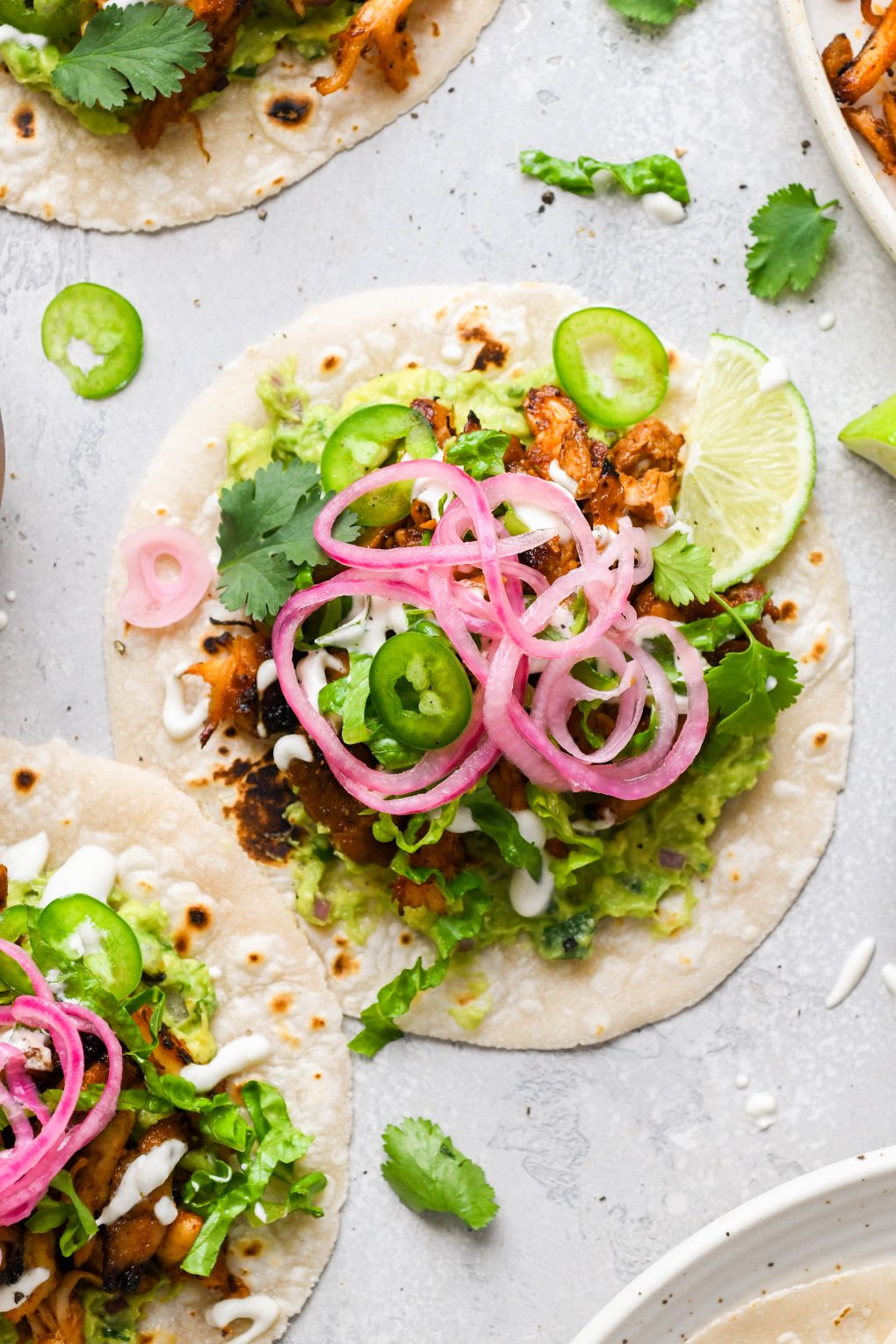 Overhead image of a few flat laid tacos on a light colored surface. They are shredded chicken tacos filled with shredded romaine, sliced jalapeno, dairy free sour cream, guacamole, cilantro, and pickled red onions.