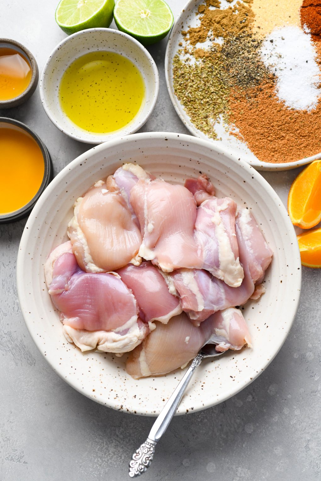 Overhead shot of the ingredients for sheet pan shredded chicken tacos - a bowl of raw boneless skinless chicken thighs, various dried spices, olive oil, orange juice, and lime juice.