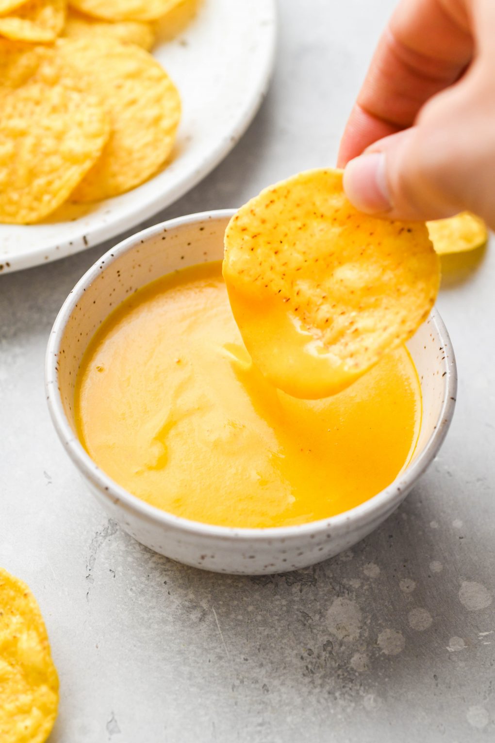 A hand dipping a round tortilla chip into a small bowl of velvety vegan cheese sauce, next to a shallow bowl of tortilla chips - on a light colored surface.