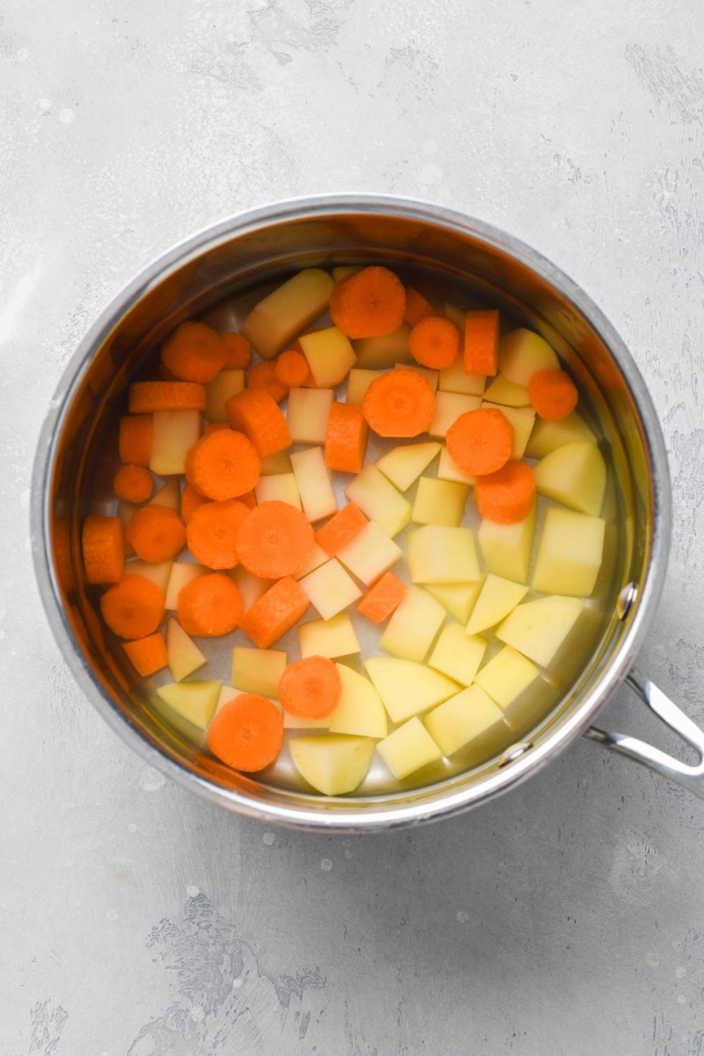 Image of cut potatoes and carrots in a small sauce pan filled with water. On a light colored background.