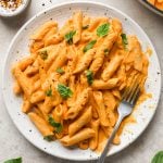 A plate of penne pasta in a creamy orange sauce, topped with fresh basil and red pepper flakes, with a fork resting on the side.