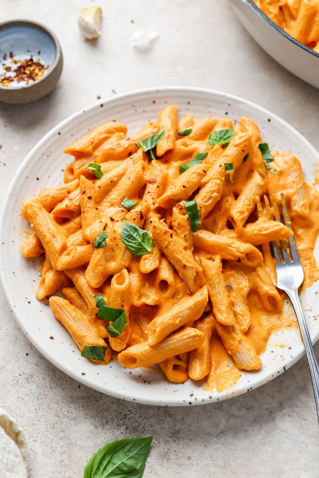 45 degree angle shot of super creamy vegan vodka pasta topped with fresh basil on an off white speckled plate on a light brown background. 