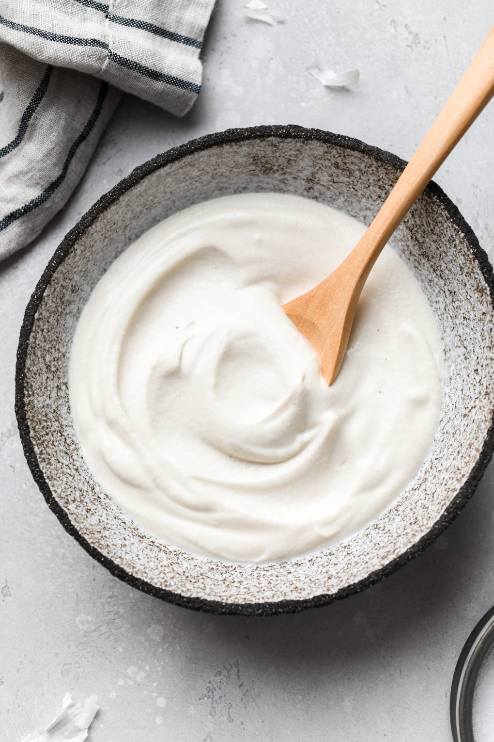Overhead shot of a small rustic looking bowl filled with creamy vegan cashew based sour cream with a wooden spoon tucked into it. On a light grey background next to a folded striped dish towel.