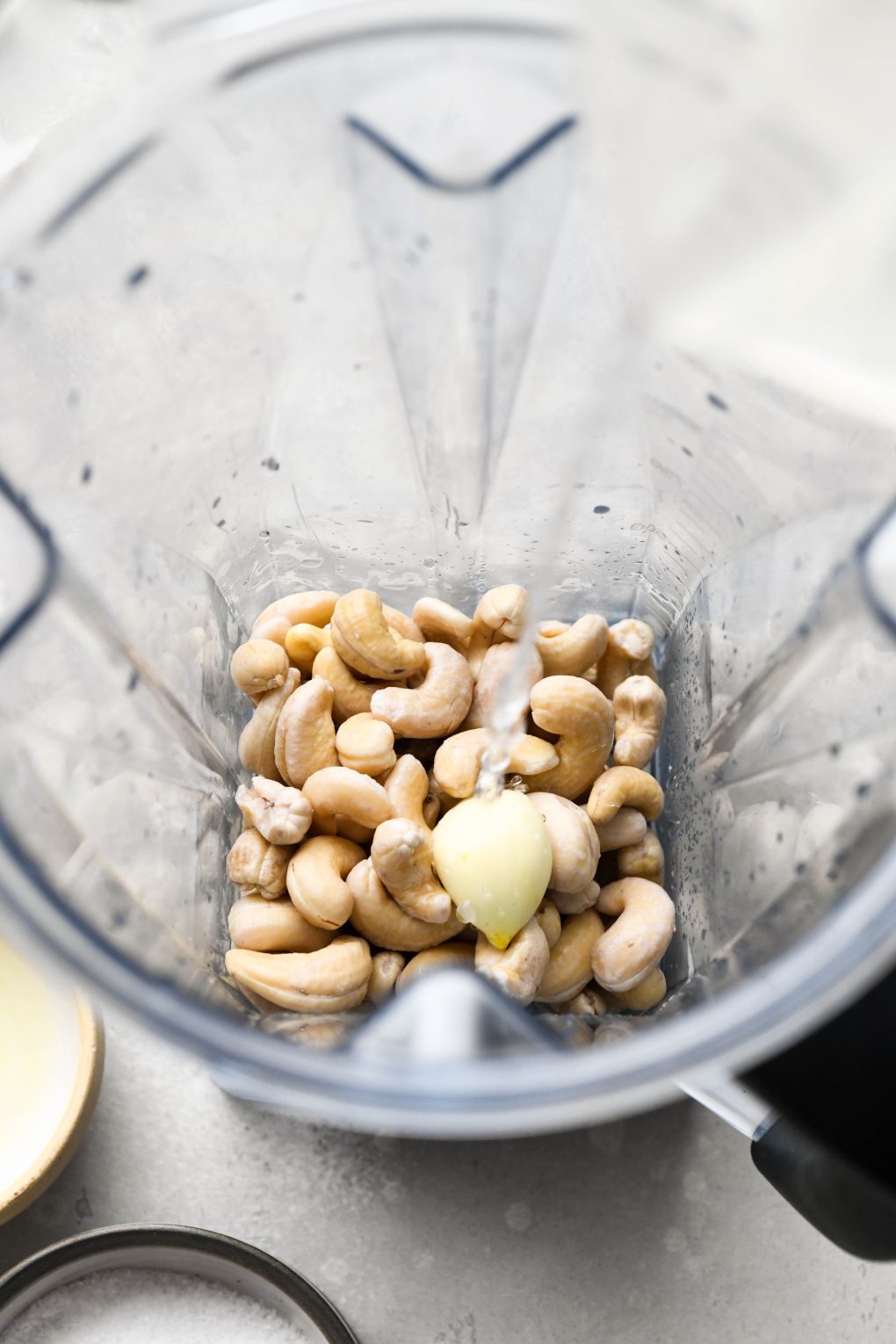 Image of water pouring into a blender container with raw cashews, garlic, and kosher salt.