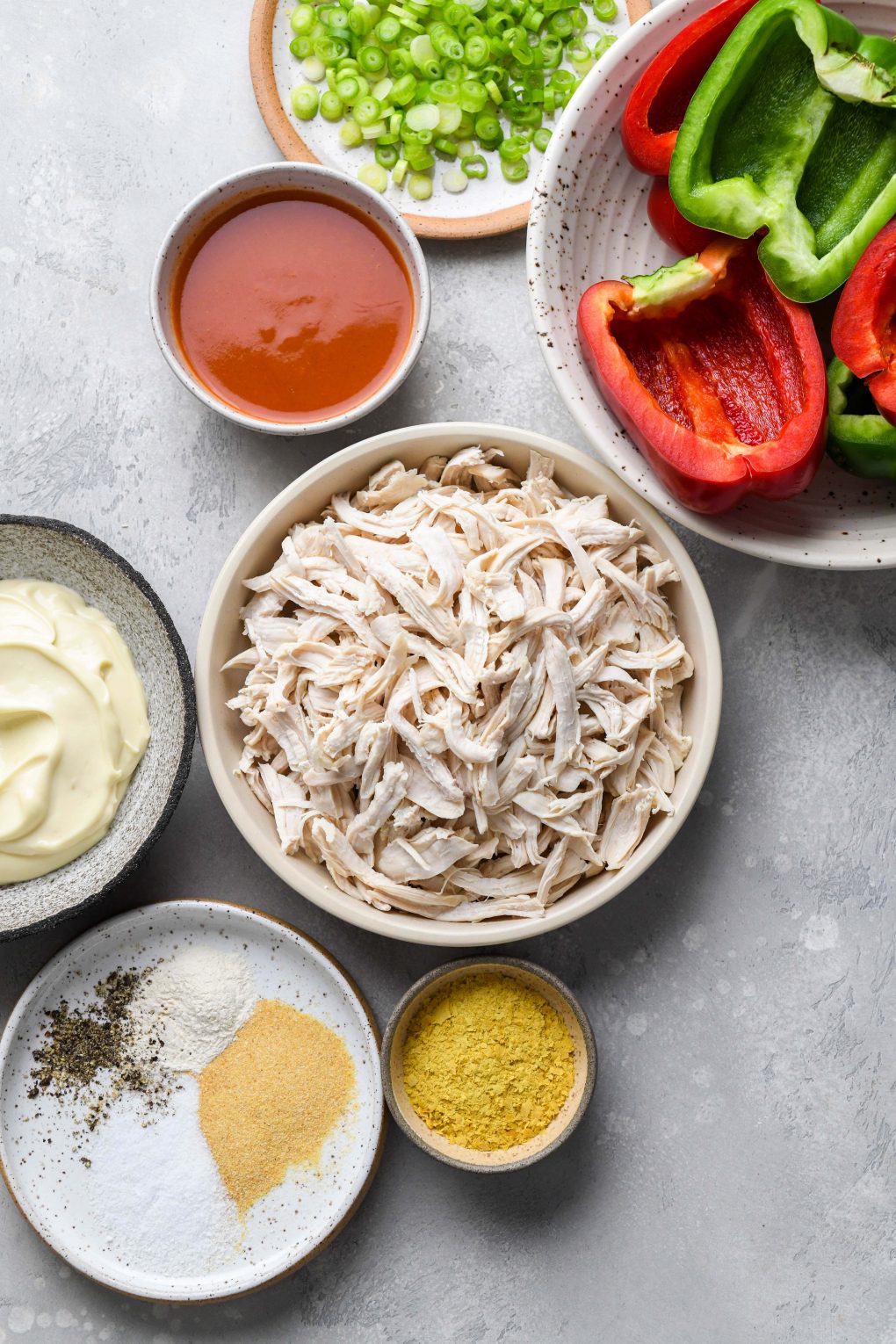 An image of ingredients used to make buffalo chicken stuffed peppers in various bowls and plates, on a light grey colored background. Shredded chicken, cut and de-seeded bell peppers, hot sauce, mayonnaise, spices, herbs, and green onions. 