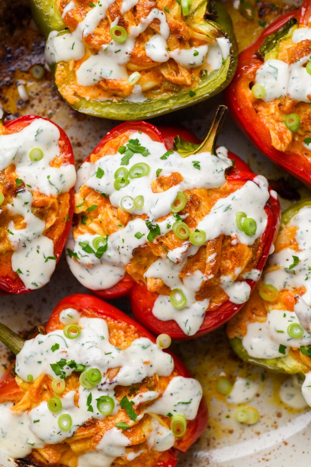 Overhead shot of colorful buffalo chicken stuffed bell peppers in an enameled cast iron skillet. Drizzled with dairy free ranch and topped with fresh herbs and thinly sliced green onion.
