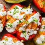 Halved red and green bell peppers stuffed with a shredded buffalo chicken filling, topped with a creamy white ranch sauce, chopped green onions, and herbs, arranged closely together on a baking dish.