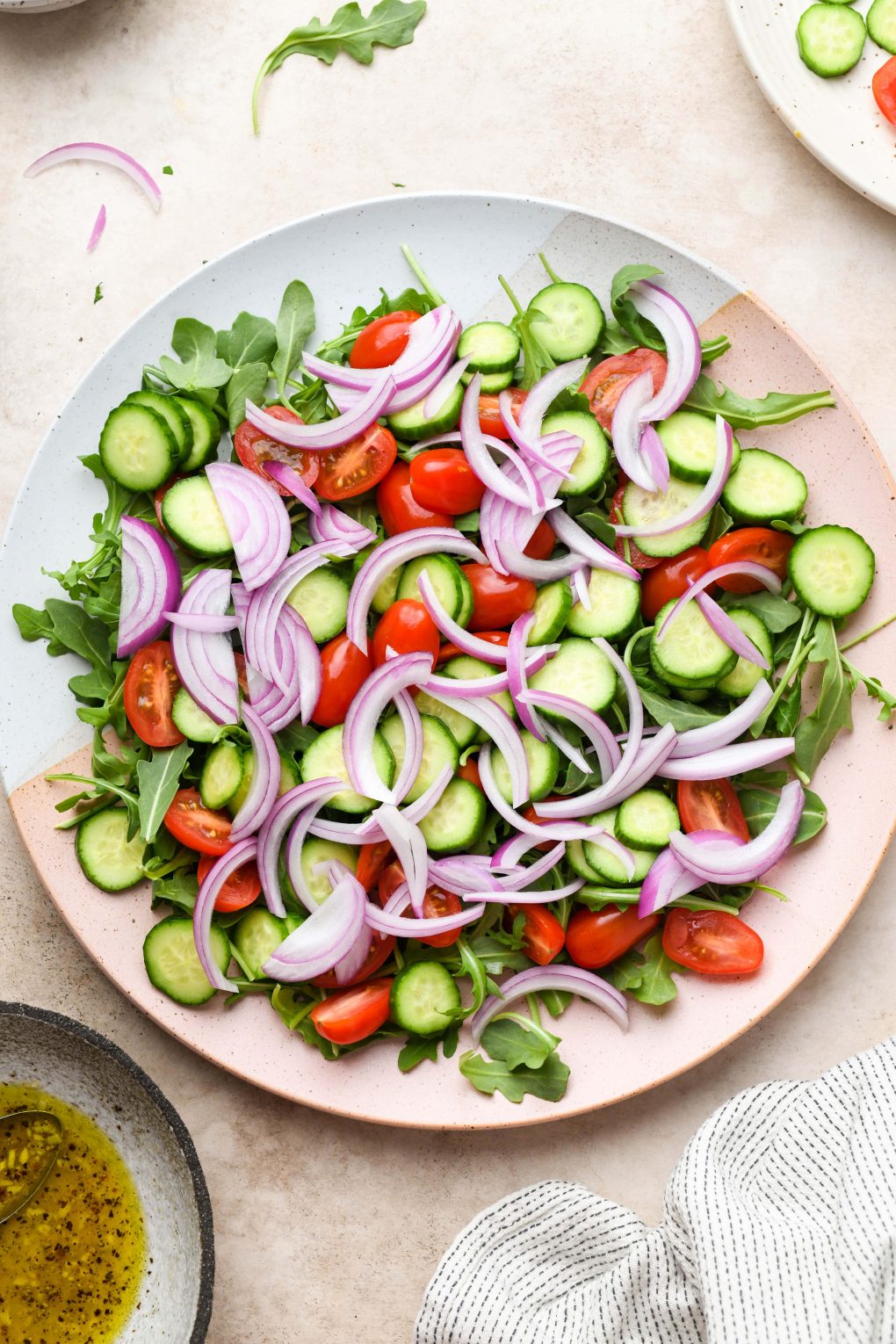 Image of large platter with arugula, cut cucumber, tomatoes, and thinly sliced red onion. On a light beige background.
