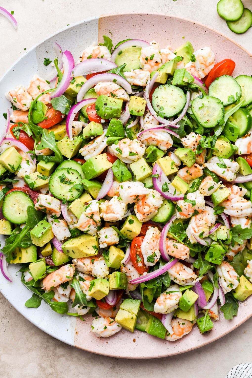 Overhead shot of a fresh and colorful shrimp and avocado salad on a pale pink and beige platter. Salad is made of arugula, sliced cucumber, cherry tomatoes, thinly sliced red onion, avocado, chopped shrimp, and fresh parsley. 