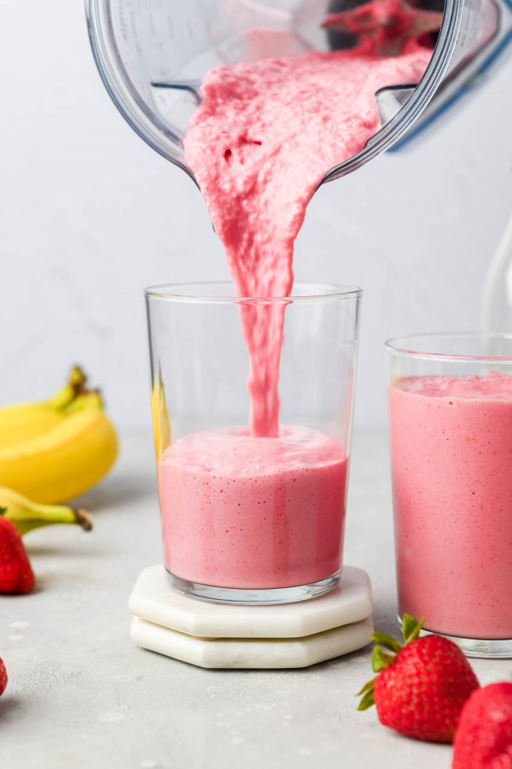 Straight on image of a blender pouring a thick strawberry banana smoothie into a glass cup. Next to a cup that's already filled with smoothie, on a light colored background.