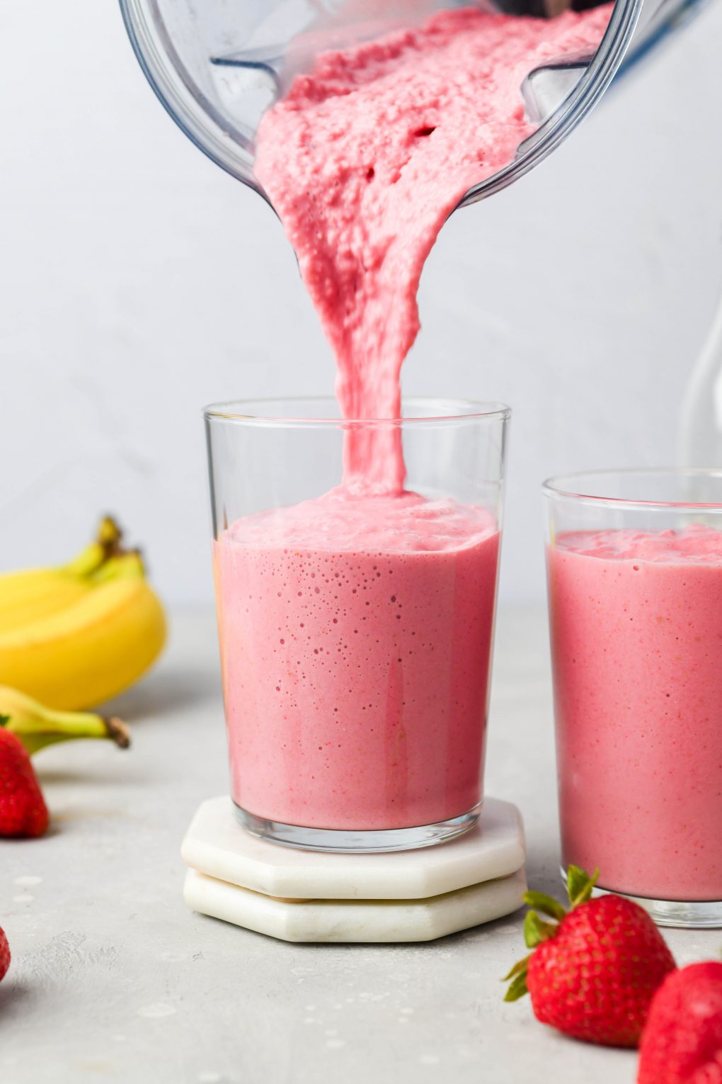 Straight on image of a blender pouring a thick strawberry banana smoothie into a glass cup. Next to a cup that's already filled with smoothie, on a light colored background.