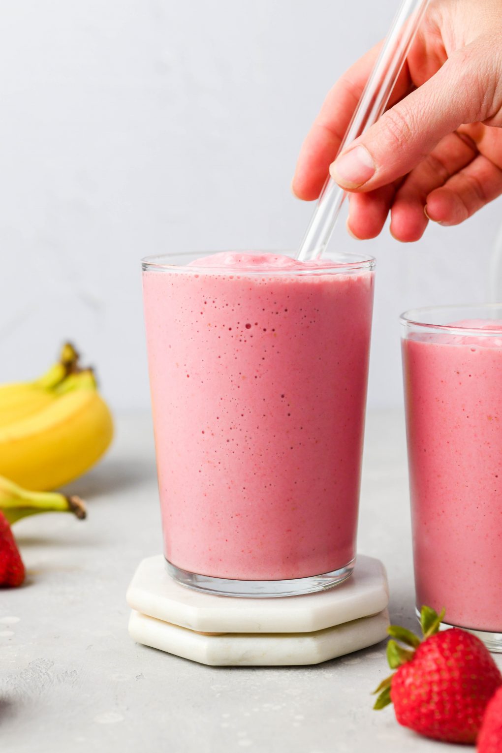 A hand reaching into the frame to place a glass straw into a glass filled with strawberry banana smoothie. On a light colored background next to some fresh strawberries and a small bunch of bananas.