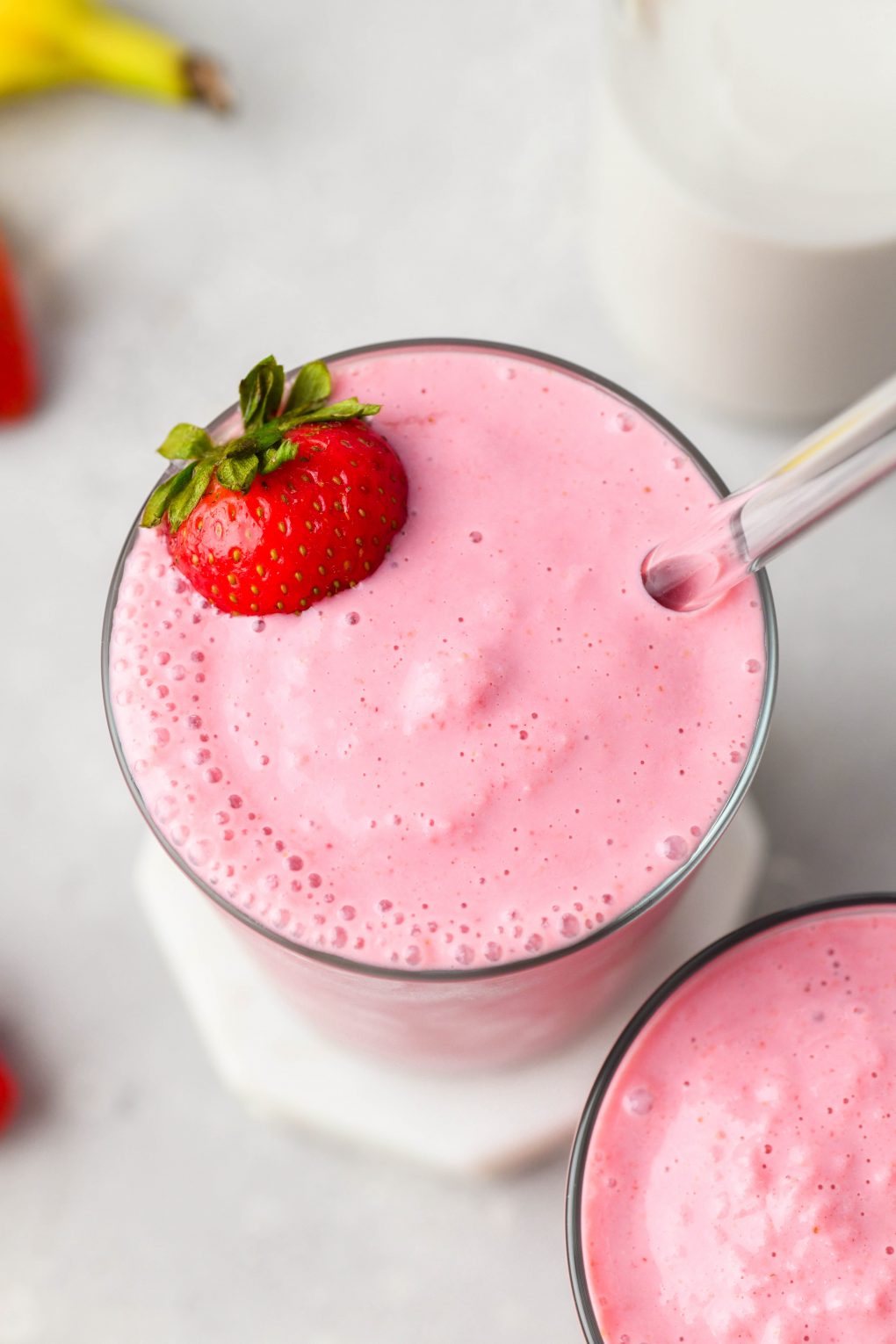 45 degree angle close up image of a strawberry banana smoothie in a glass cup. Topped with a fresh strawberry. On a light colored background. 