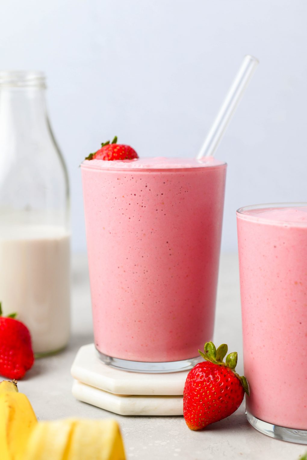 Straight on shot of 2 side by side glasses of strawberry banana smoothie. On a light colored background next to a glass jar of dairy free milk, some fresh strawberries, and a banana peel. 
