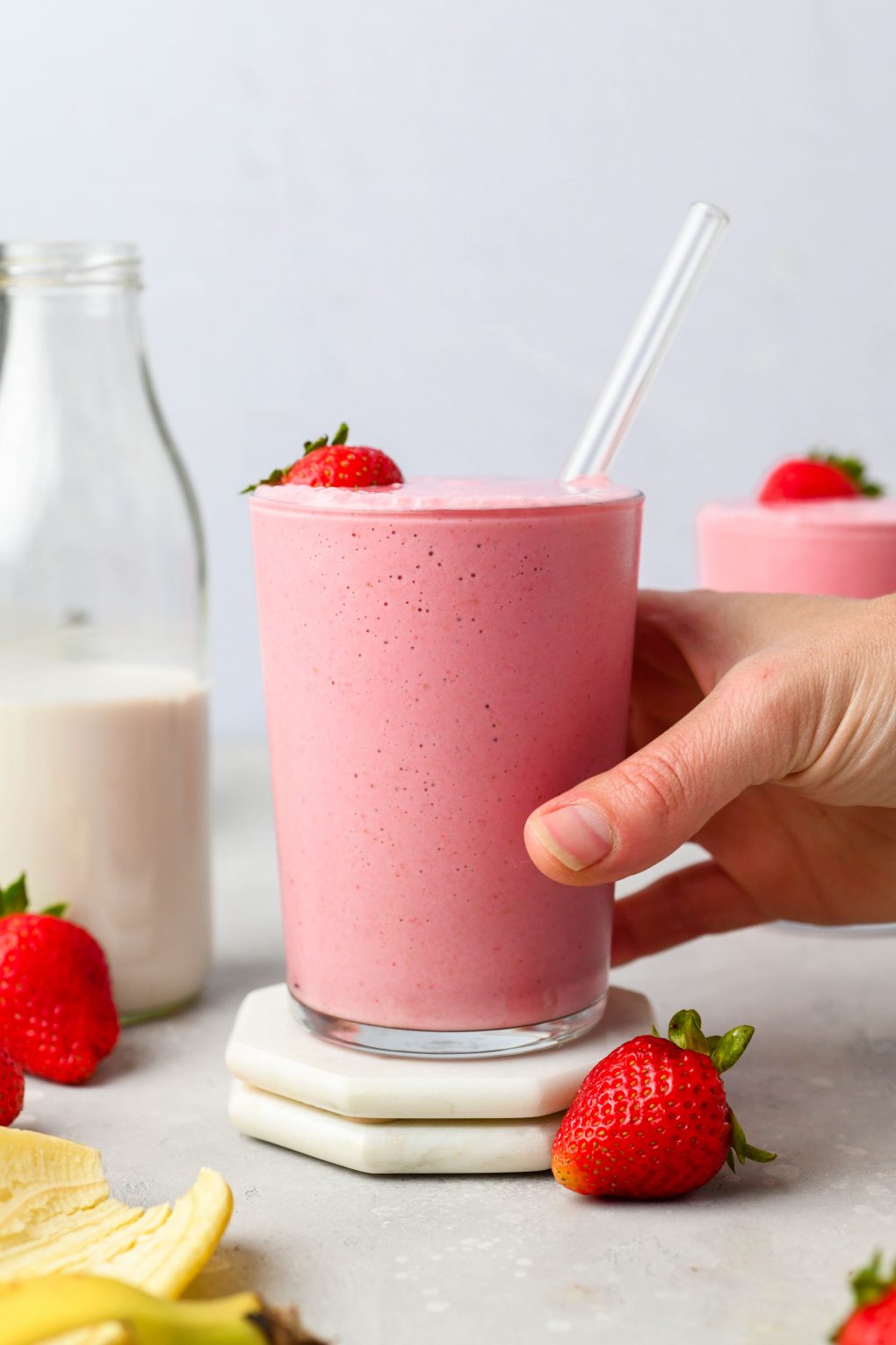 A hand reaching into the frame to grab a glass filled with a creamy strawberry banana smoothie. On a light background, next to some fresh strawberries and a glass jar of dairy free milk.