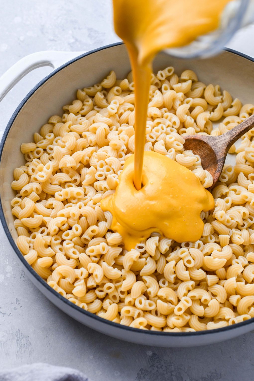 Side angle shot of creamy vegan cheese being poured into a large skillet of cooked macaroni pasta.