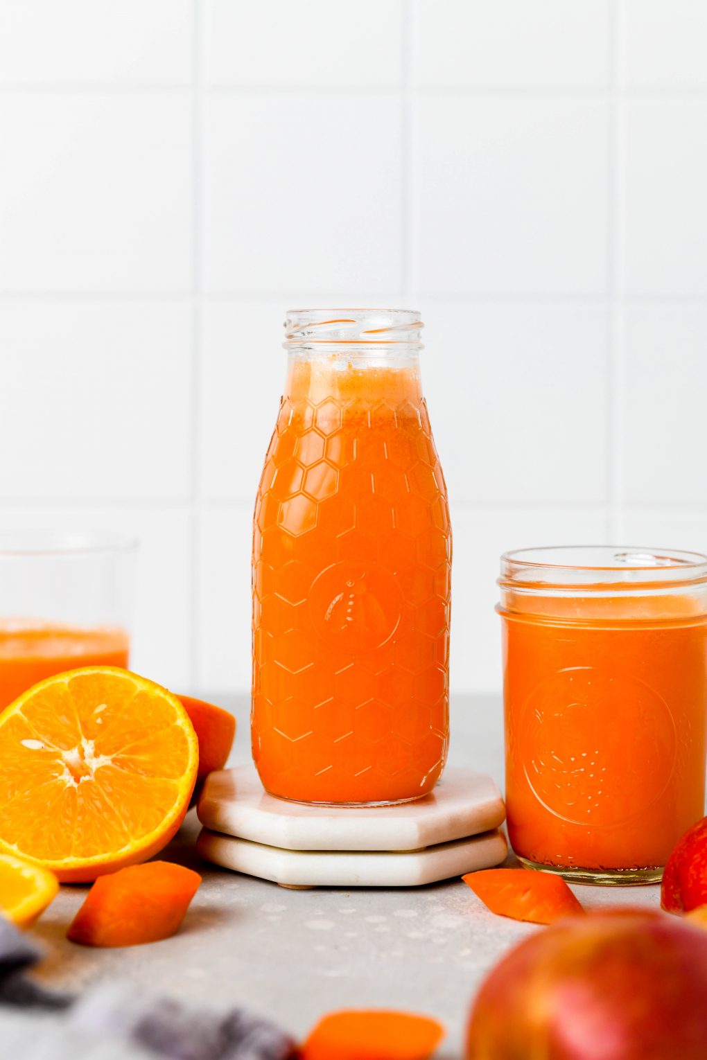 Straight on shot of a textured glass jar filled to the brim with bright orange carrot, apple, and orange juice with ginger. Next to some cut oranges, and carrot pieces, on a light colored background.