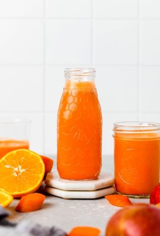 A glass bottle and a jar filled with bright orange juice sit on a table, surrounded by fresh orange halves and carrot pieces. The background is a white tiled wall.