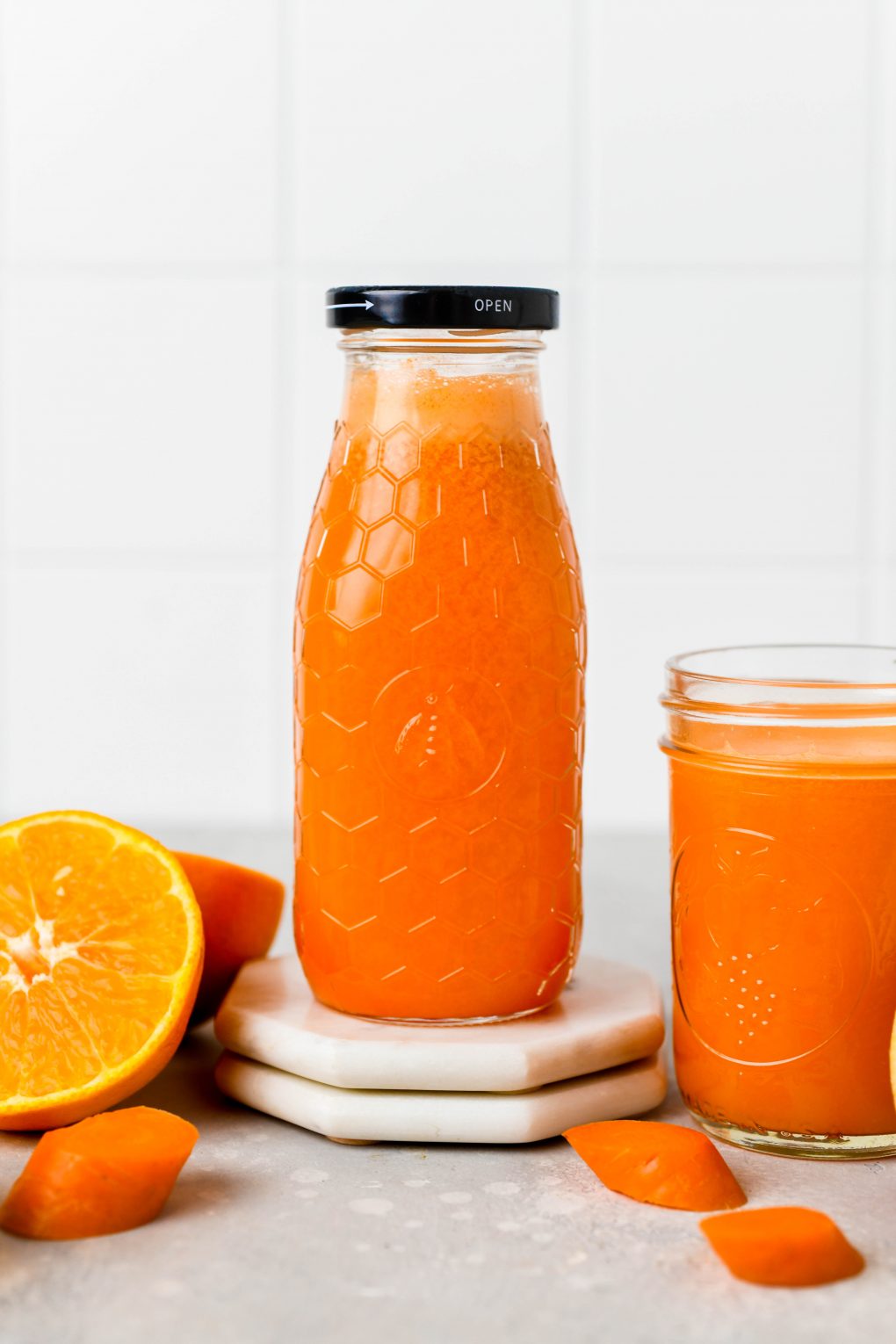 Straight on shot of a textured glass jar filled to the brim with bright orange carrot, apple, and orange juice with ginger. Next to some cut oranges, and carrot pieces, on a light colored background.