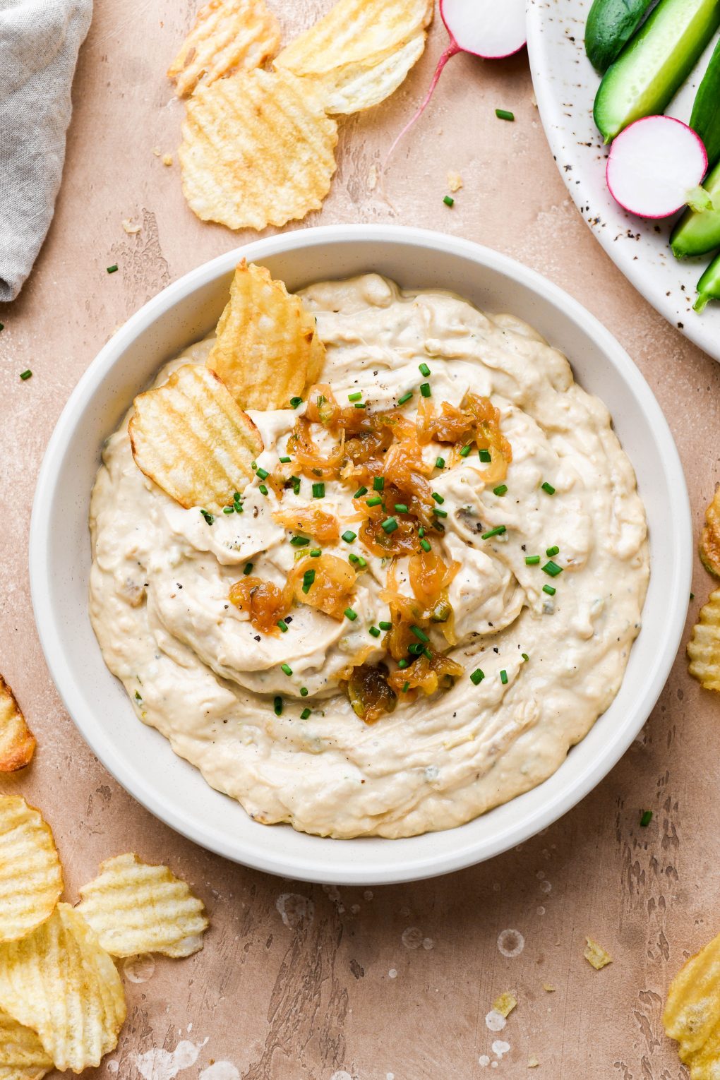 Close up overhead shot of a shallow cream colored bowl filled with super thick and creamy dairy free french onion dip. Dip is topped with caramelized onions and chives. On a light brown background surrounded by scattered ruffle potato chips and a plate of cut veggies.