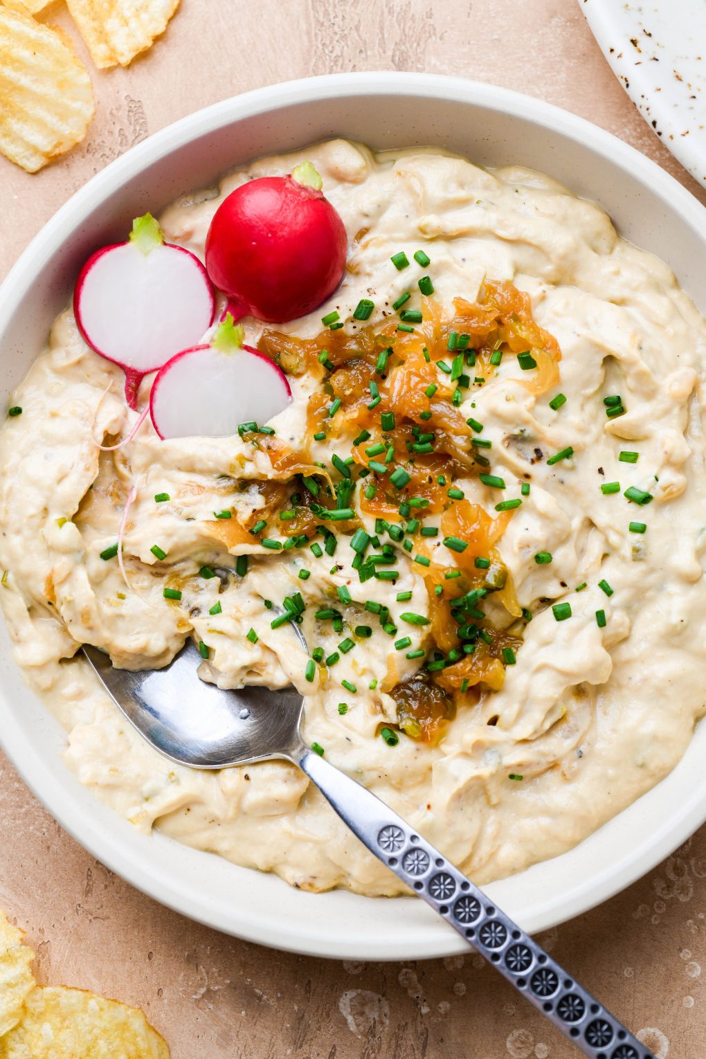 Close up overhead shot of a bowl of dairy free french onion dip, topped with caramelized onions, snipped chives, and some cut radishes tucked into the side of the bowl.