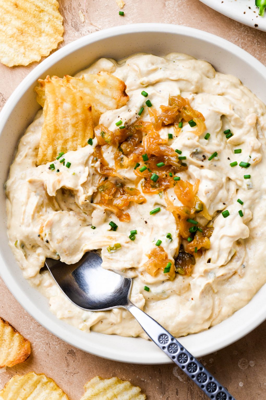 Close up overhead shot of a shallow cream colored bowl filled with super thick and creamy dairy free french onion dip. Dip is topped with caramelized onions and chives. On a light brown background surrounded by scattered ruffle potato chips. 