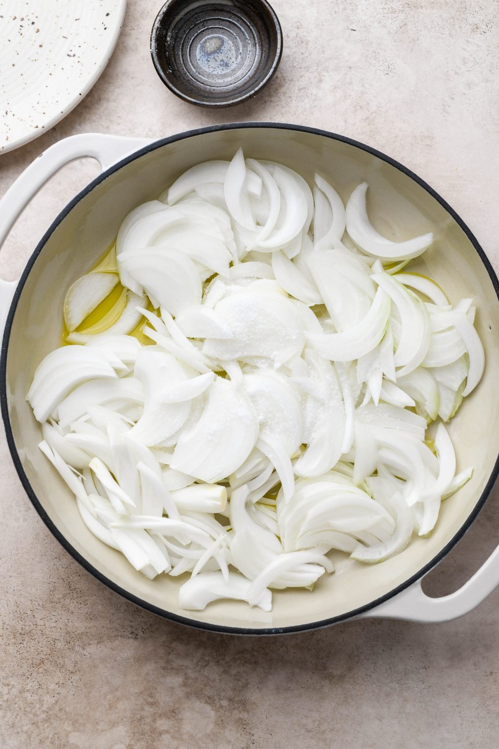 Image of a large cream colored ceramic coated skillet with olive oil, thinly sliced onion, and salt. On a light brown background. 