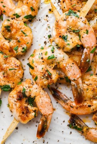 Close up image of grilled shrimp on wooden skewers. On a light colored speckled plate topped with chopped parsley.