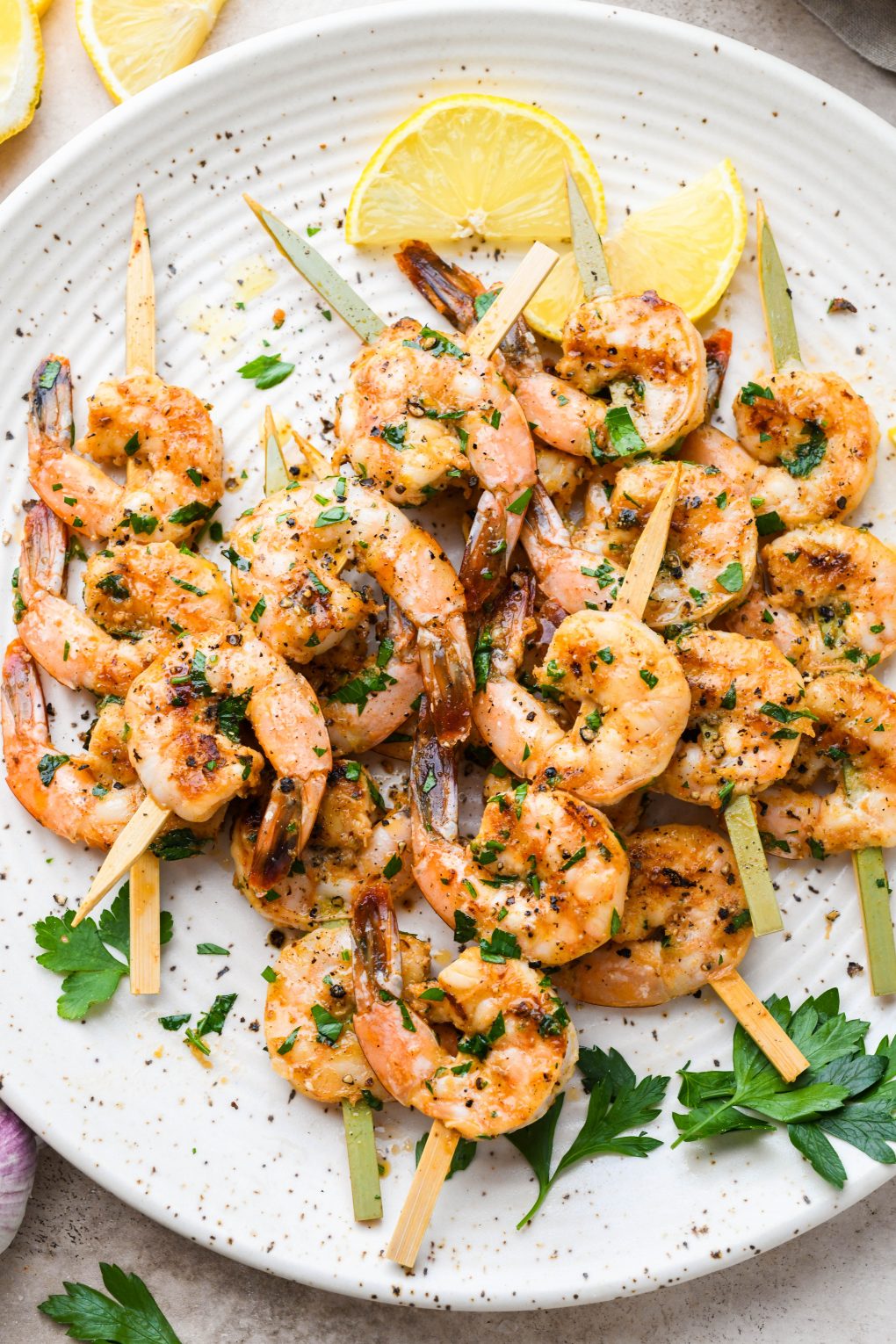 Overhead image of grilled shrimp on wooden skewers. On a light colored speckled plate topped with chopped parsley and next to some lemon wedges.