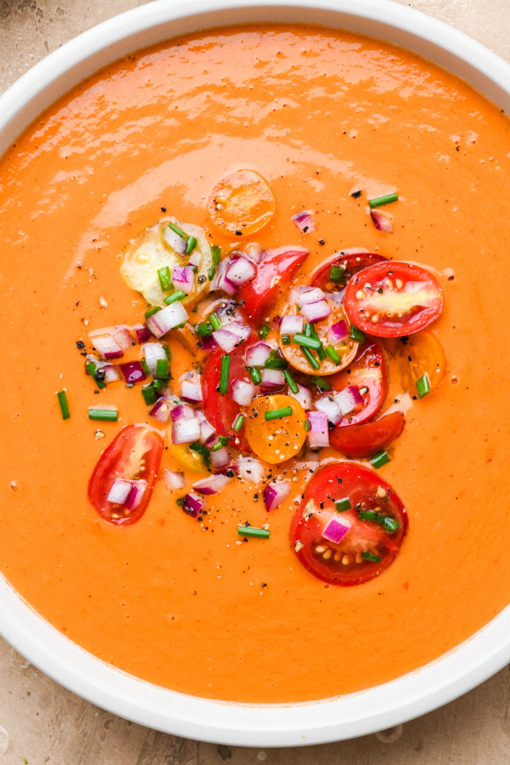 A large shallow bowl of bright orange healthy gazpacho. Topped with a cherry tomato salad and snipped fresh chives. On a light brown textured background.