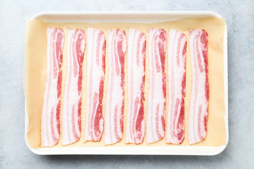 Raw bacon slices on a white baking sheet lined with parchment paper