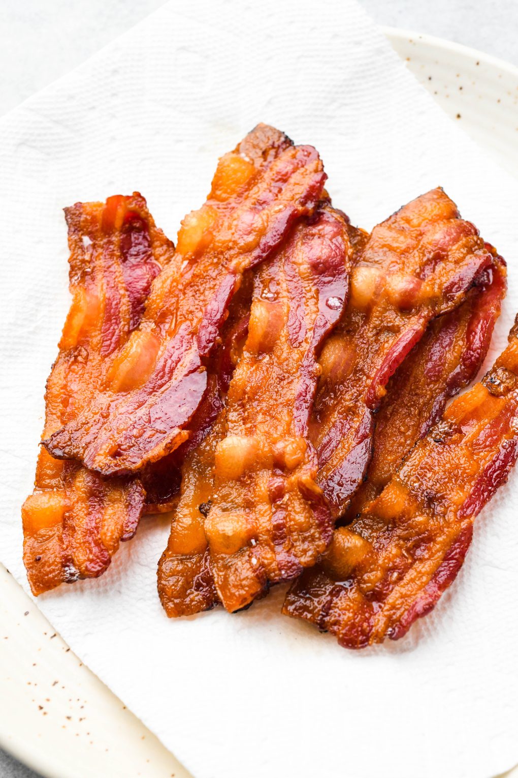 Crispy strips of bacon on paper towels, on top of a white speckled plate.