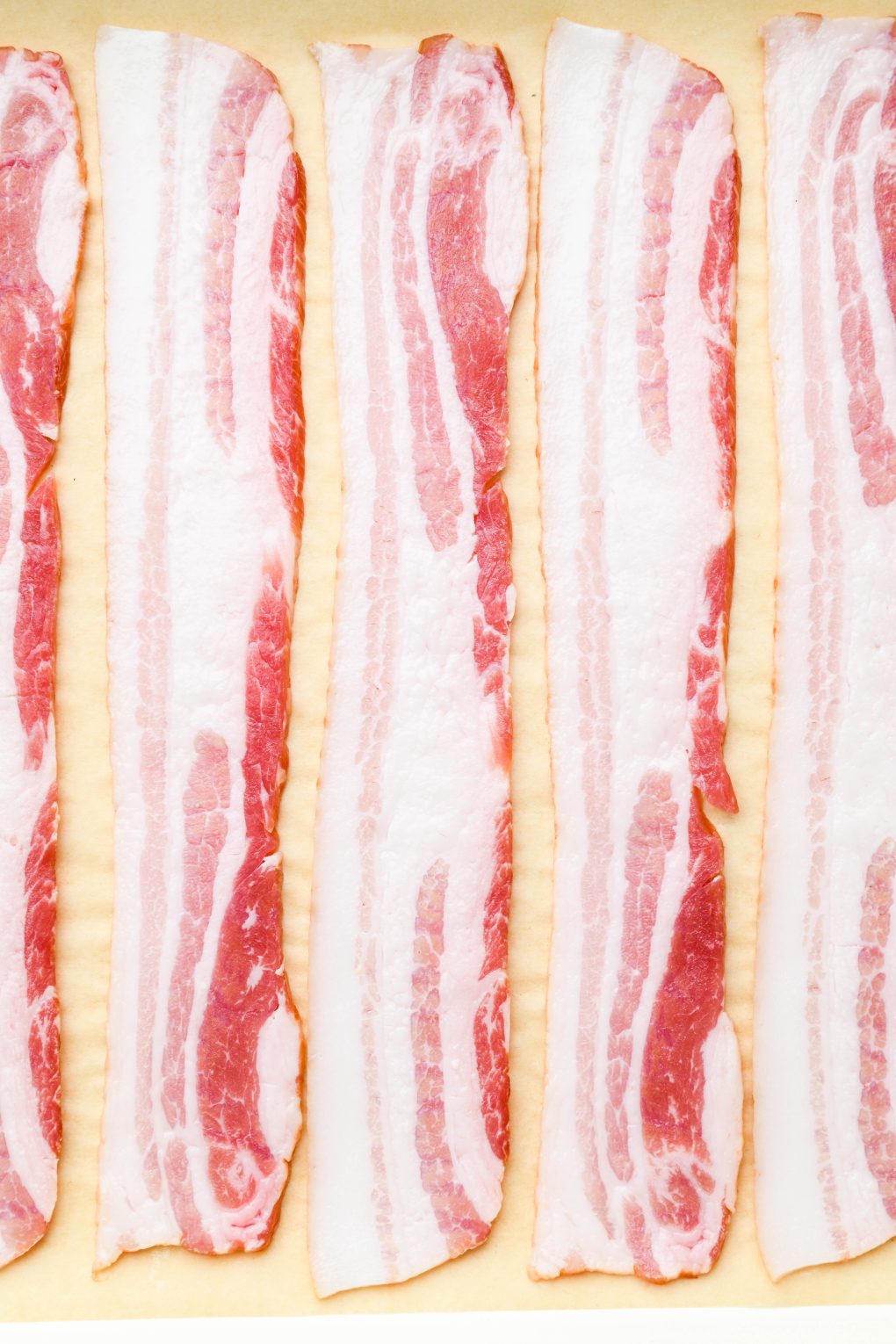 Close up of raw bacon slices on a white baking sheet lined with parchment paper