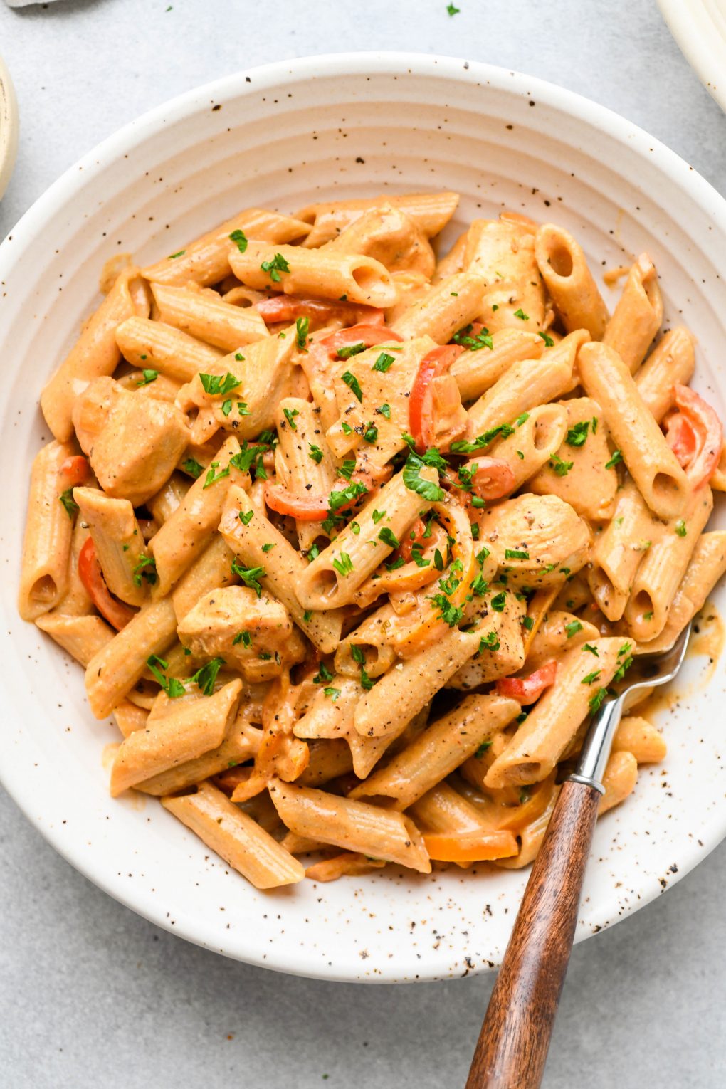 Large speckled shallow bowl filled with creamy cajun chicken penne pasta. Topped with a sprinkle of parsley and a wooden handled fork tucked into the side of the bowl. 