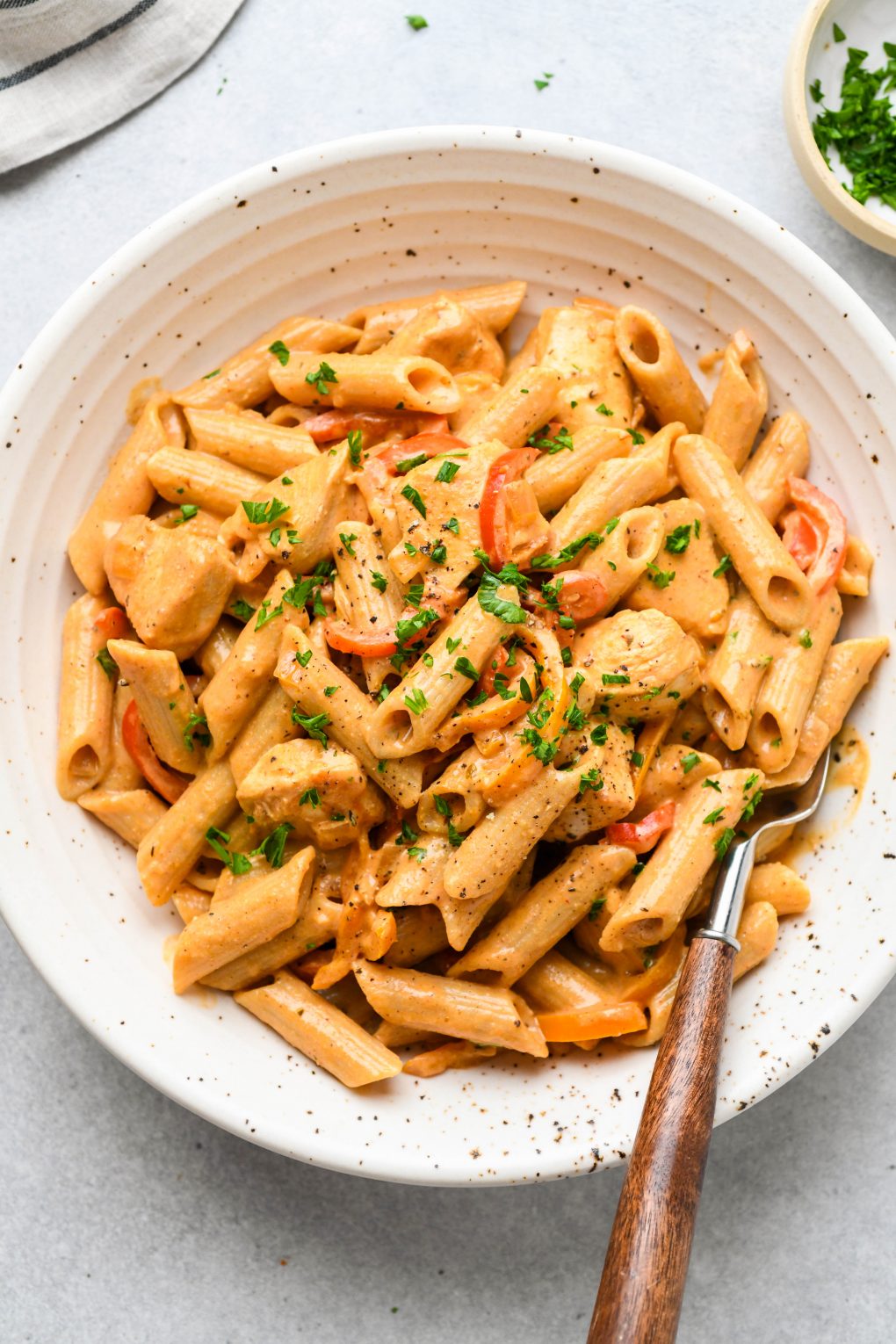 Large speckled shallow bowl filled with creamy cajun chicken penne pasta. Topped with a sprinkle of parsley and a wooden handled fork tucked into the side of the bowl. 
