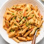 A bowl of creamy penne pasta with sliced red bell peppers and sprinkled chopped parsley, served with a fork on the side.