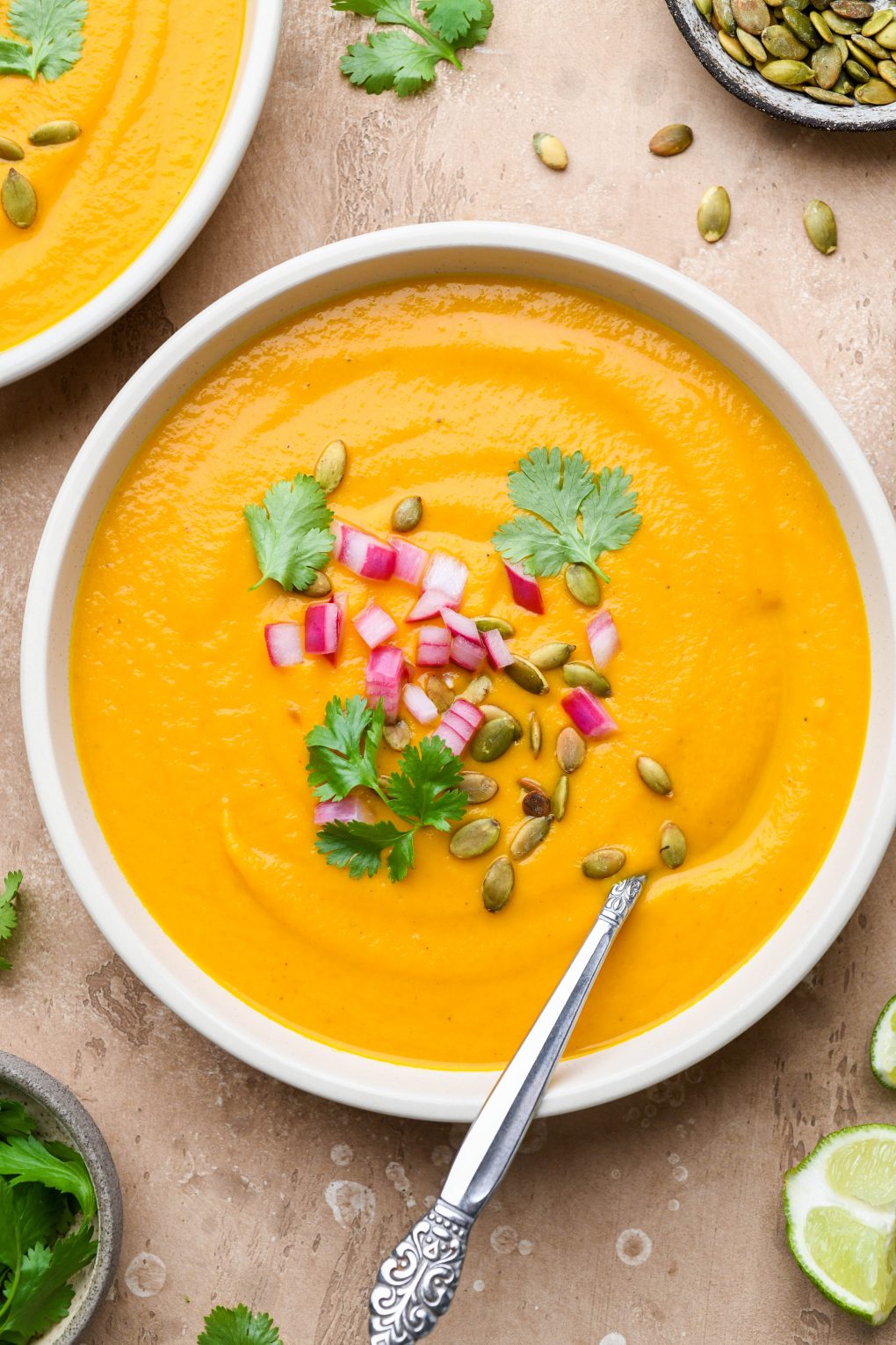Large wide bowl of bright orange creamy carrot soup on a light brown background. Soup is topped with fresh cilantro leaves, chopped pickled red onions, and pumpkin seeds.