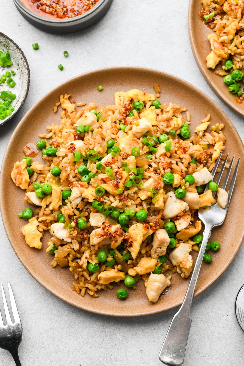 A plate of gluten free chicken fried rice garnished with green onions and chili oil, next to a second plate, a small dish of green onions, a bowl of chili oil, and two forks.