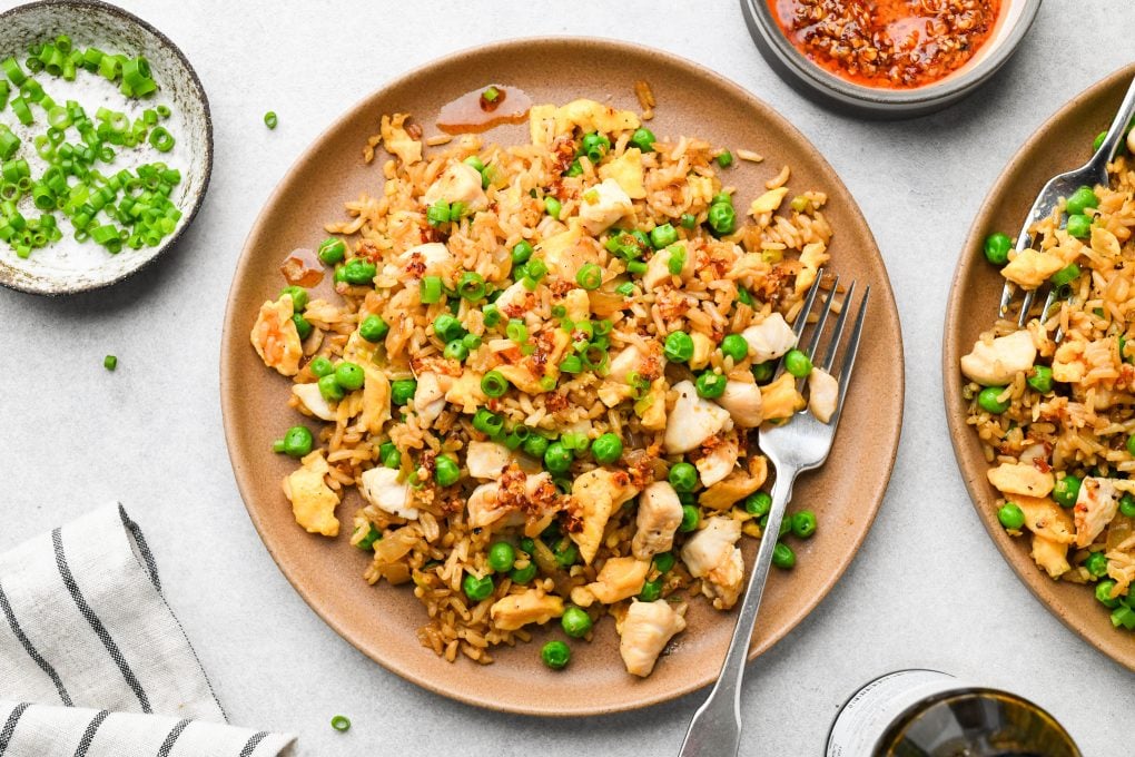 A plate of gluten free chicken fried rice garnished with green onions and chili oil, next to a second plate, a small dish of green onions, and a bowl of chili oil. 