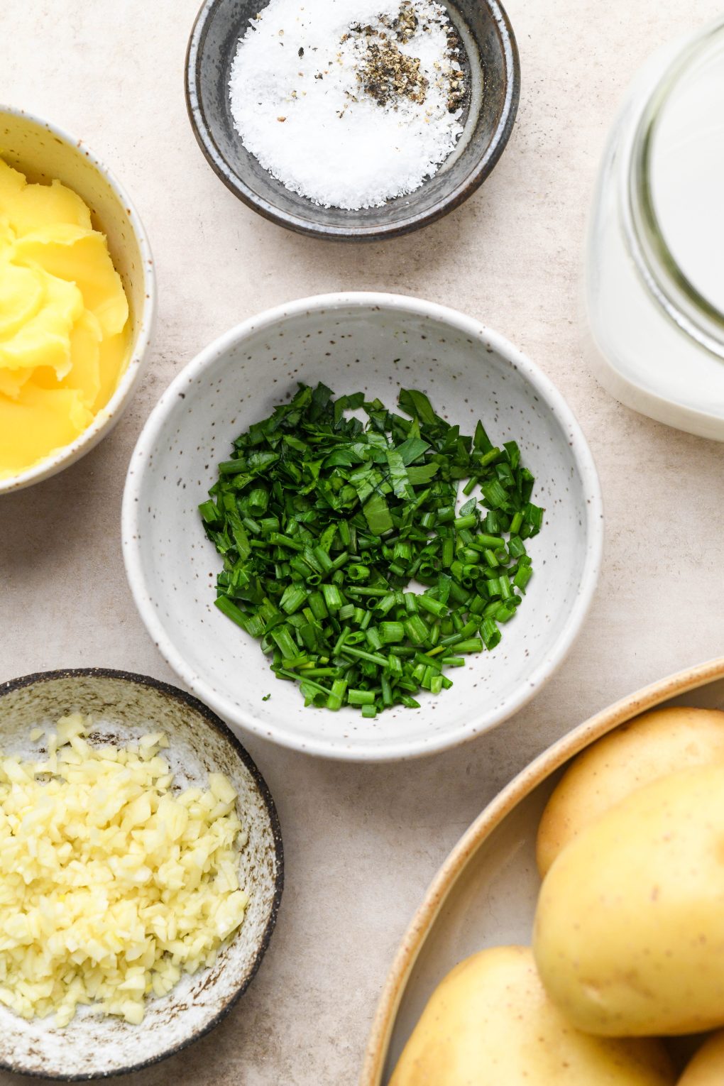 Ingredients for Whole30 mashed potatoes - overhead view of a small bowl of herbs, ghee, salt and pepper, chopped garlic, and a jar of dairy free milk.