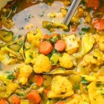 A pot of vibrant vegetable soup with chunks of carrot, cauliflower, cabbage, and fresh herbs, with a ladle lifting some of the soup.