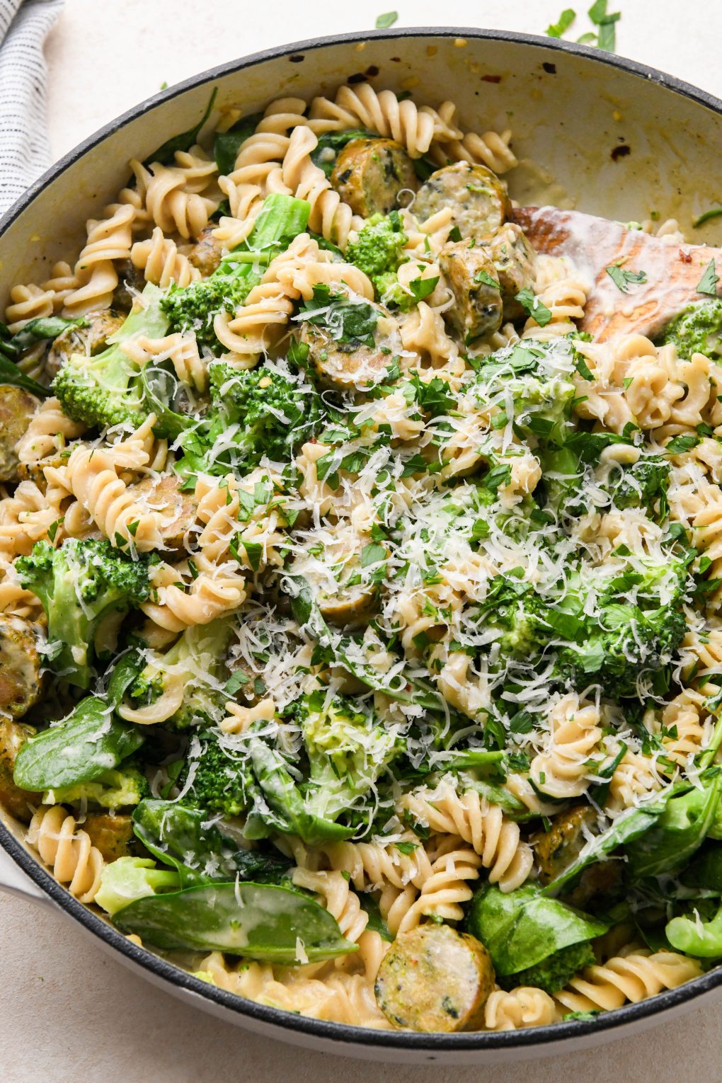 Healthy chicken sausage pasta in a ceramic skillet topped with vegan parmesan and fresh parsley on a cream colored background.