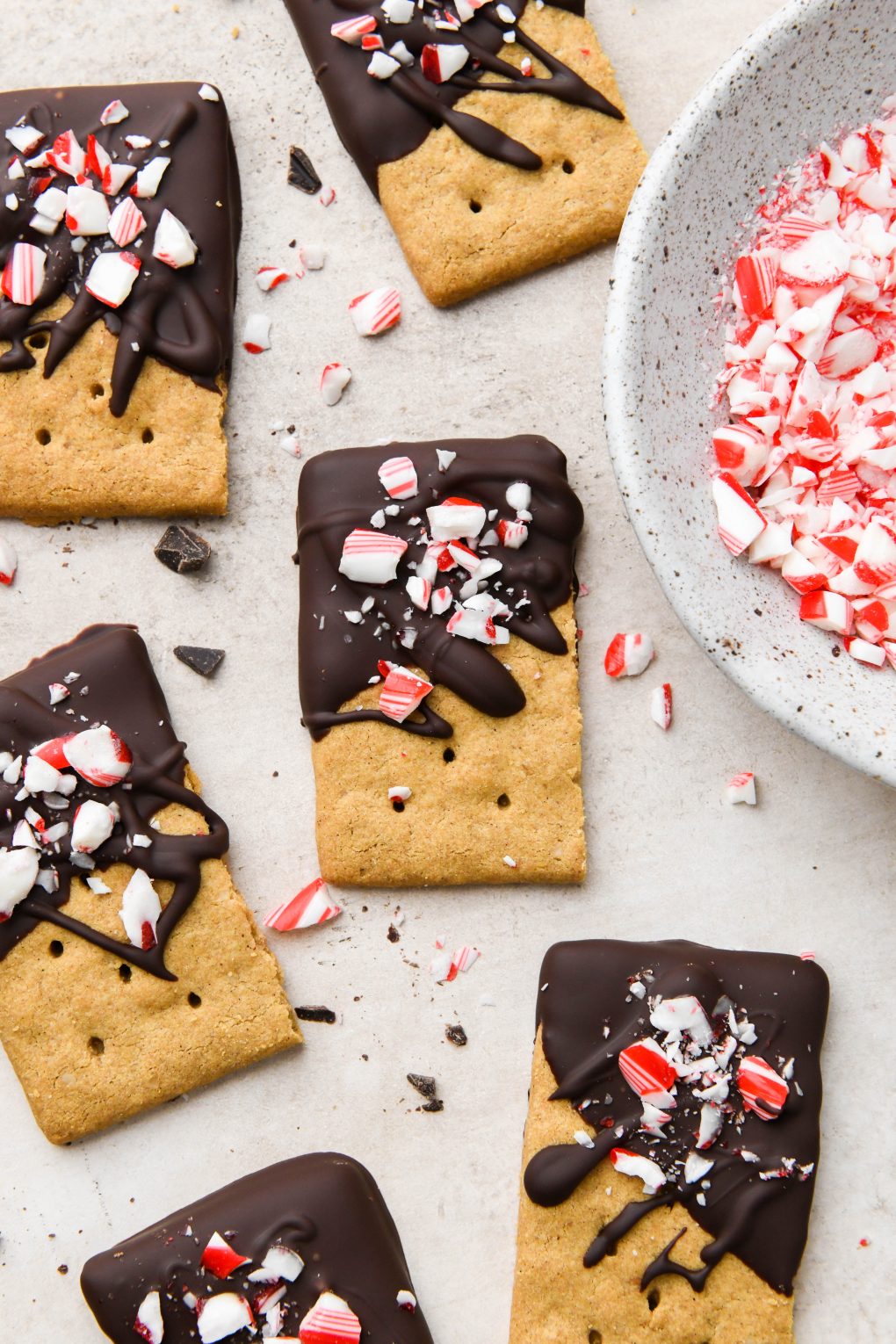 Chocolate dipped graham crackers topped with crushed candy canes on a light brown surface next to a bowl of crushed candy canes.
