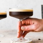 A hand holding a coupe glass filled with a dark espresso martini topped with a creamy foam, placed on round marble coasters. Coffee beans are scattered on the light surface nearby.